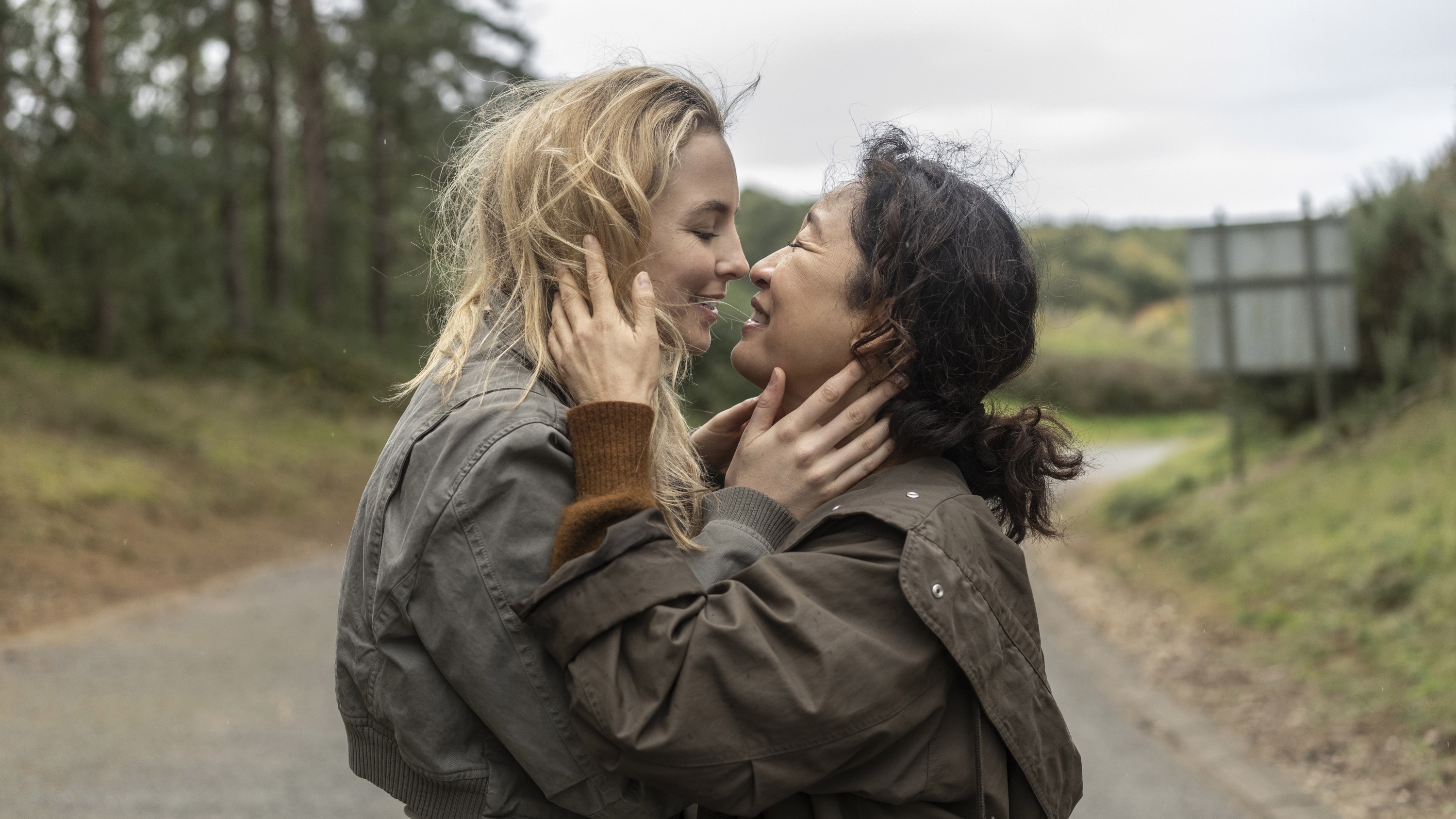 Jodie Comer and Sandra Oh embrace and smile at each other outdoors, both wearing casual jackets, in a scene from "Killing Eve."