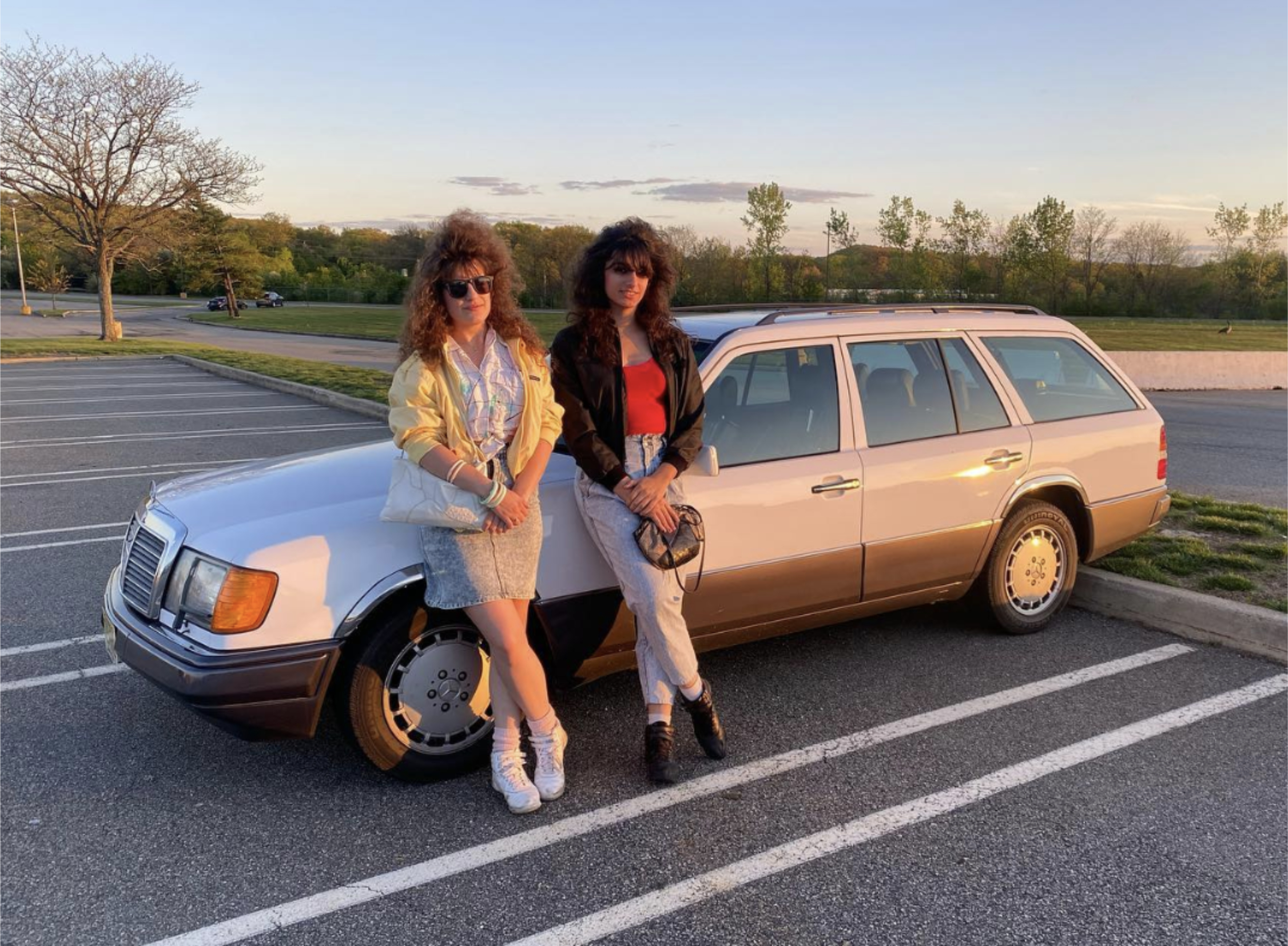 Two women with 1980s fashion, one in a yellow jacket and skirt, the other in a black jacket and jeans, pose in front of a vintage station wagon in a parking lot