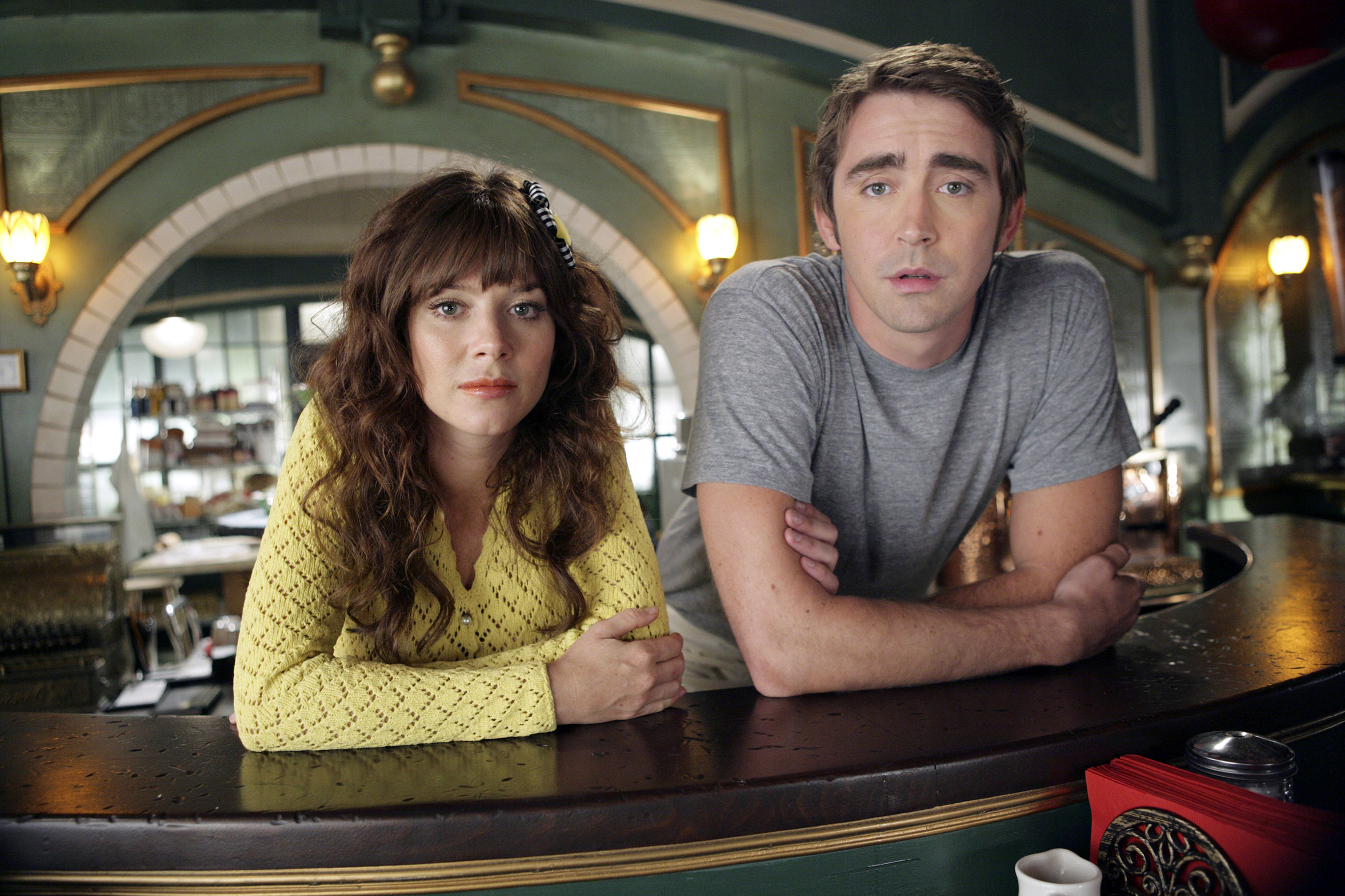 Zooey Deschanel in a patterned top and Lee Pace in a plain t-shirt lean on a counter in a retro-styled diner setting