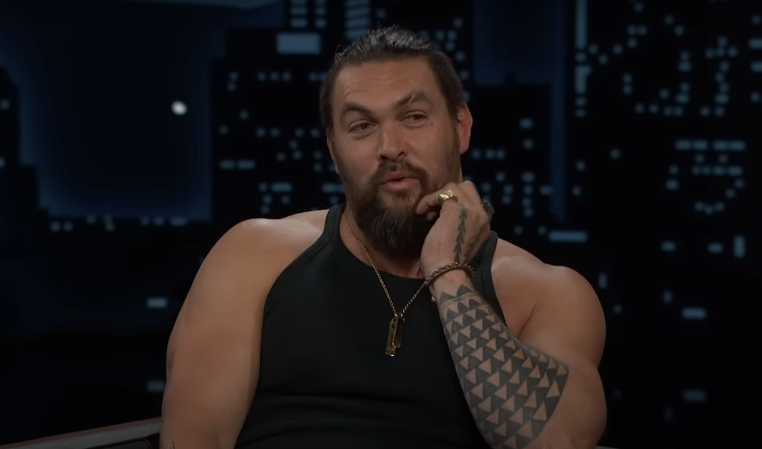 Jason Momoa, in a sleeveless tank top, showcases his arm tattoo while speaking on a nighttime talk show