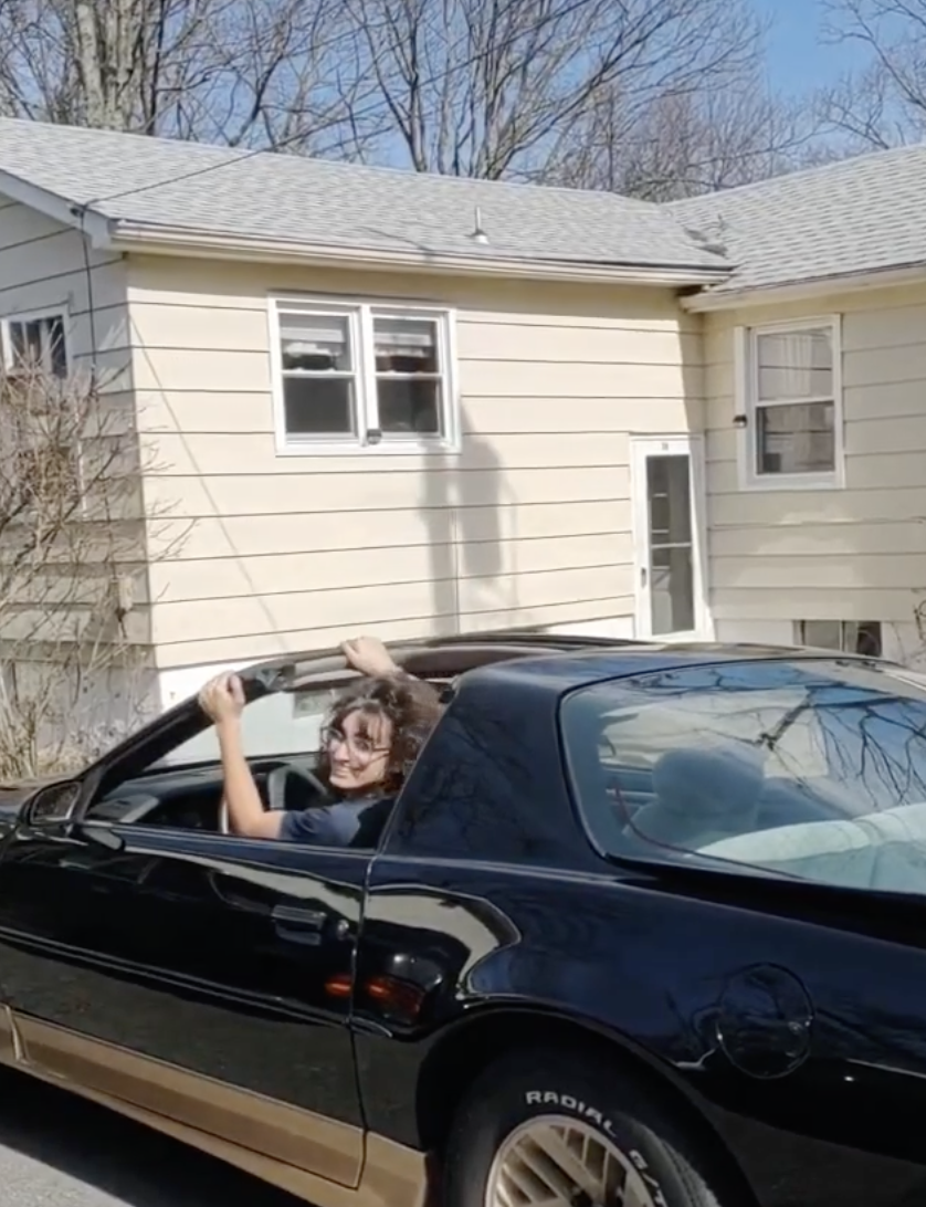 A person smiles while raising their arms up, sitting in a black convertible car in front of a beige house. The top is down, and it is daytime