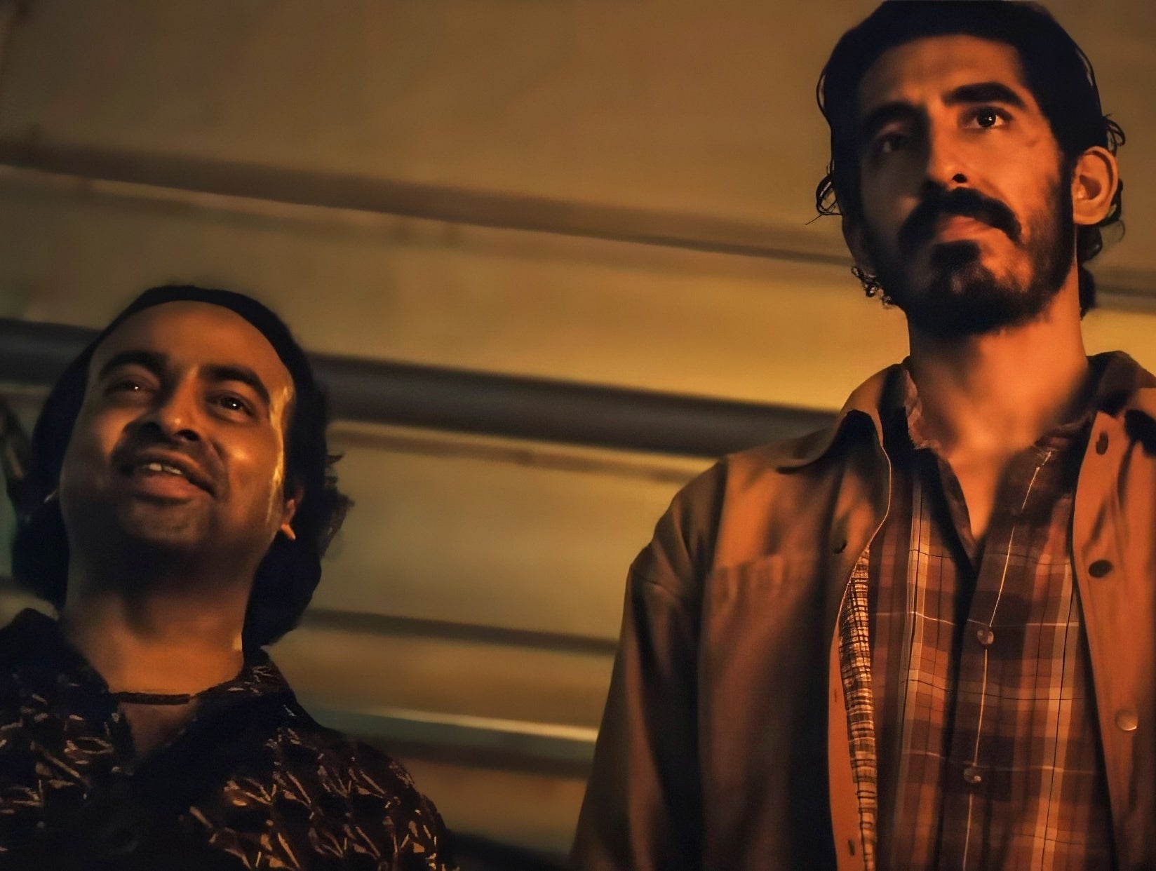 in a scene, Dev Patel in a dimly lit indoor setting, wearing a plaid shirt under a jacket