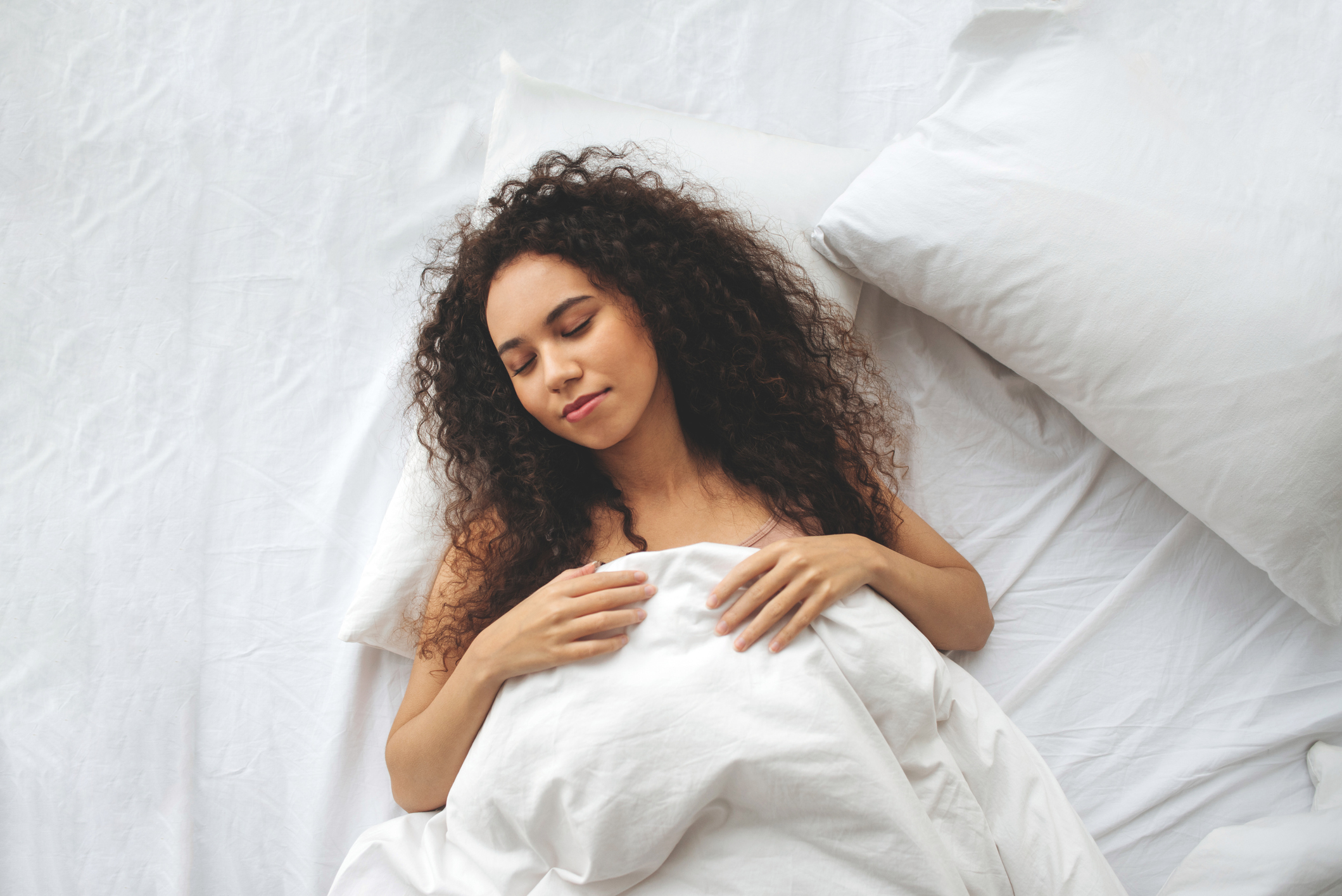 A person with curly hair lies peacefully in bed, covered by white sheets, with a soft smile on their face, portraying relaxation and contentment