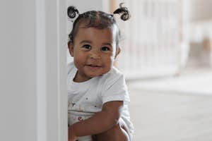 A toddler, with hair in two small pigtails, wearing a white outfit, smiles while crouching behind a door frame in a room with a crib in the background