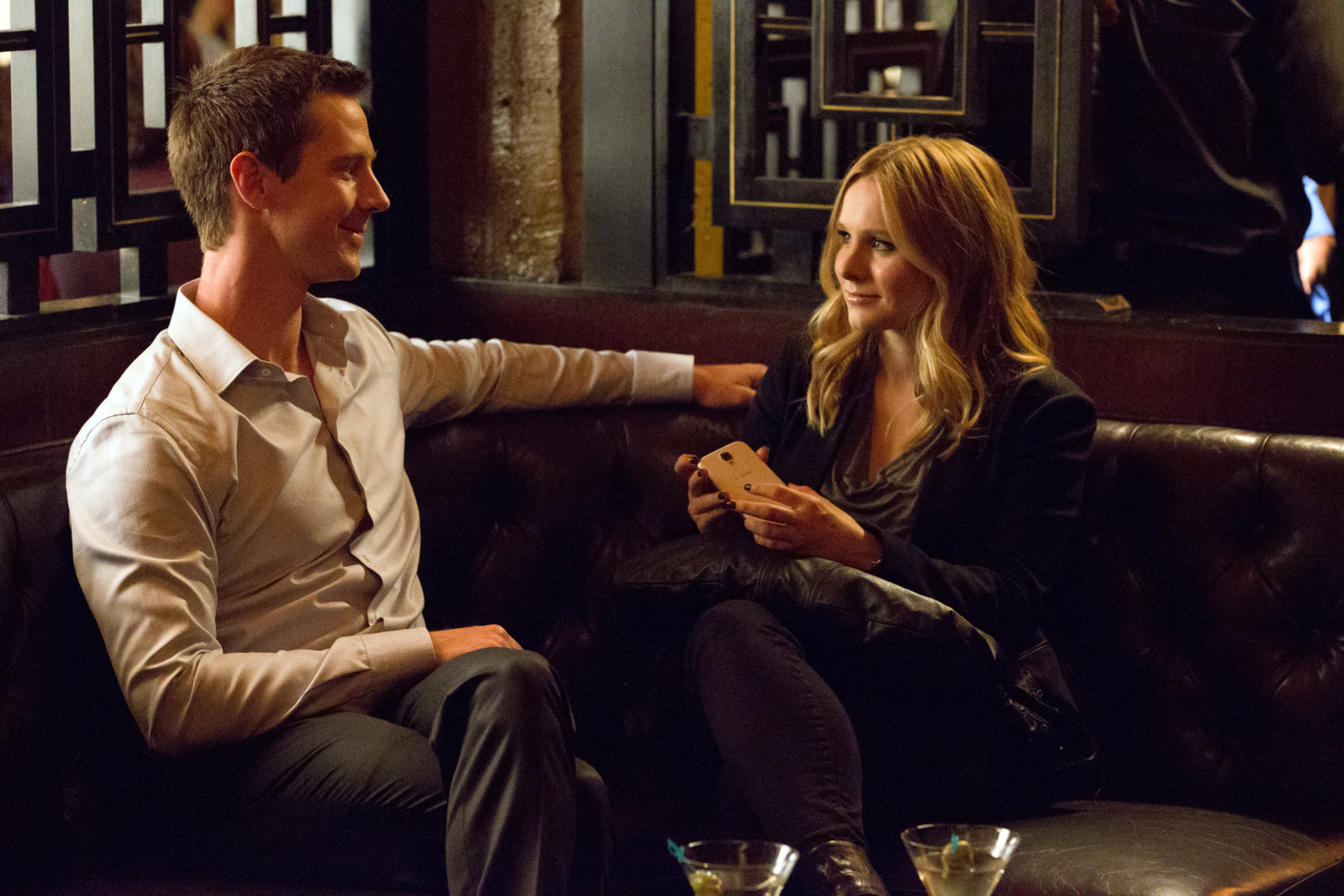 Jason Dohring and Kristen Bell seated on a couch, talking in a cozy bar setting, both dressed in casual attire