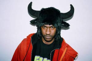 Skepta wearing a black furry hat with bull horns, a black graphic T-shirt, and an orange jacket, poses against a plain background