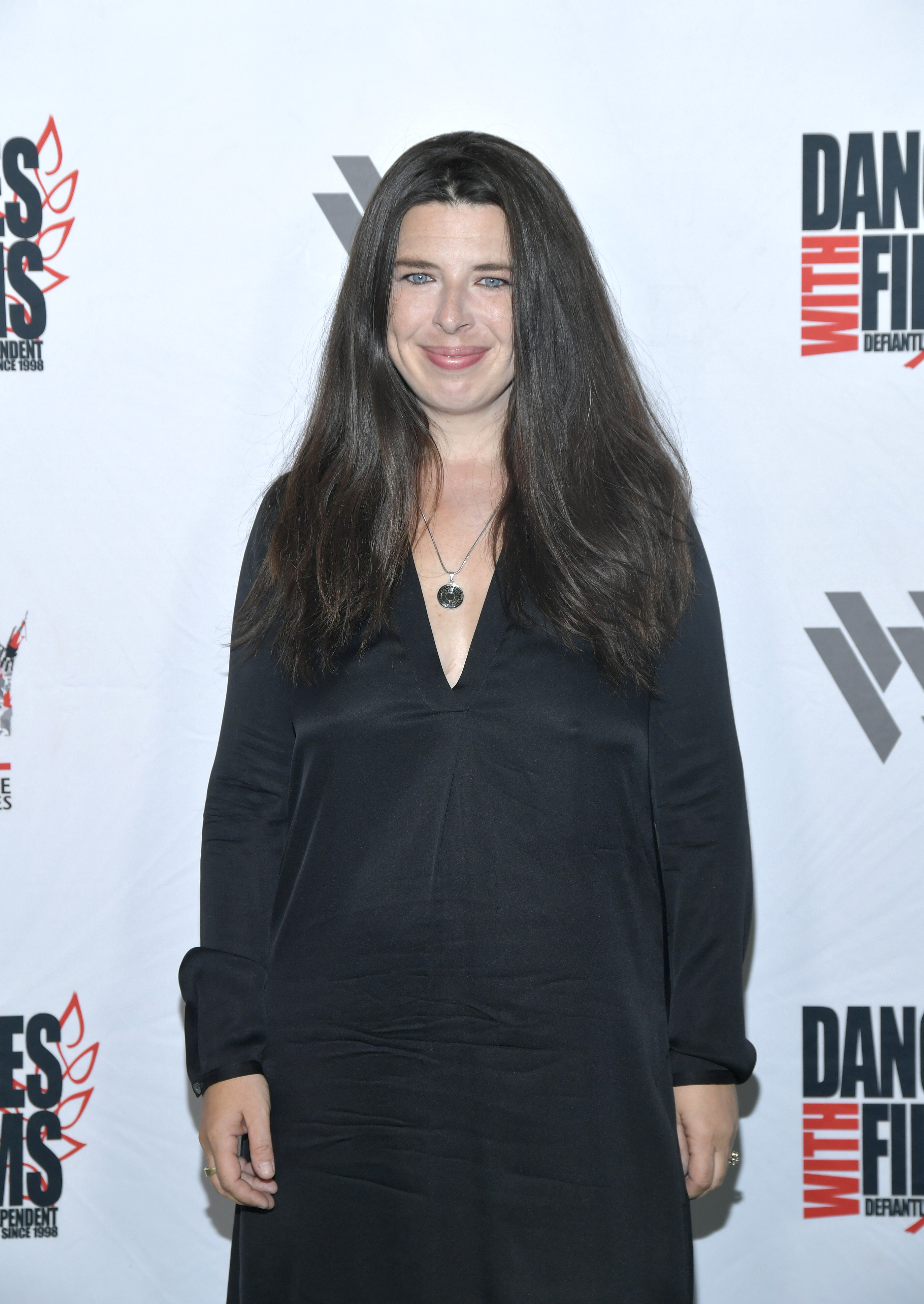 Heather Matarazzo with long dark hair stands in front of a 'Dances With Films' backdrop, wearing a black V-neck dress and a pendant necklace