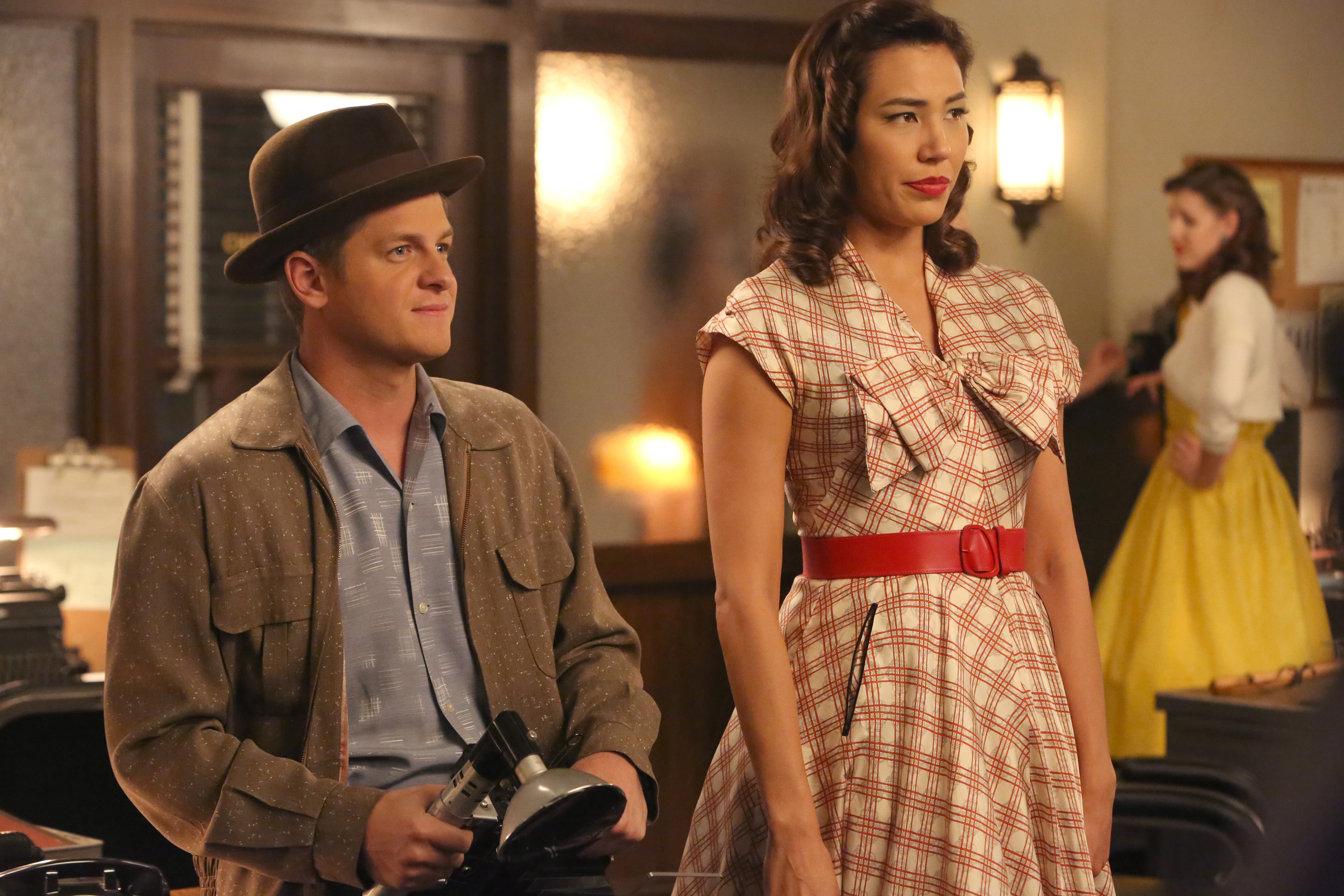 Aaron Stanford and Tamara Taylor in period attire on the set of a TV show, with Aaron holding a camera and Tamara in a dress with a red belt