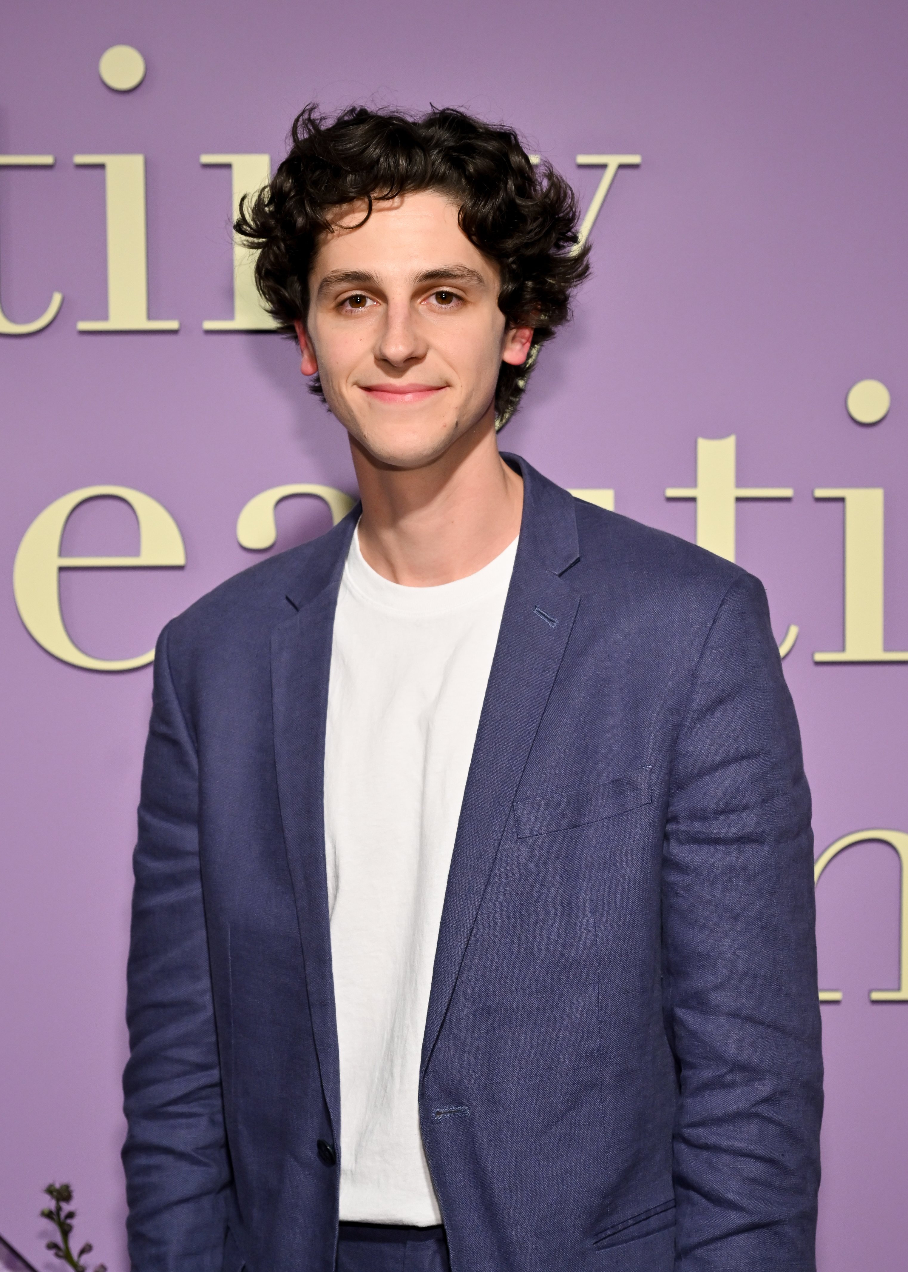 Owen Painter wearing a white t-shirt and blue suit, smiling at a TV/movie event with a purple background
