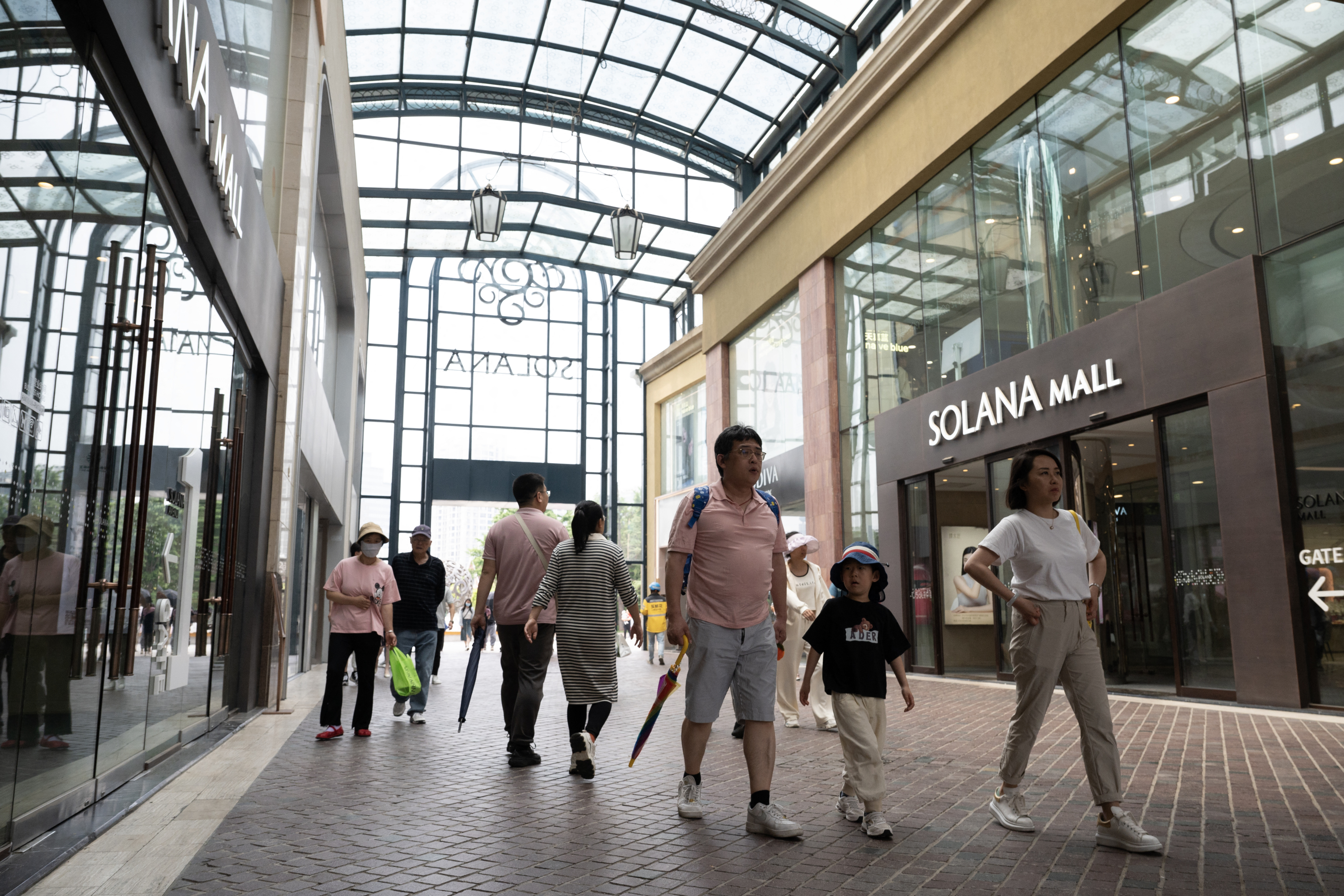 People walking inside Solana Mall, some holding umbrellas. Stores with glass fronts and a high arched ceiling are visible
