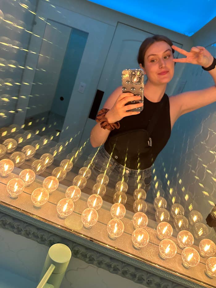 Woman in black sleeveless top and blue jeans taking a mirror selfie in a room with many small light bulbs. She is making a peace sign