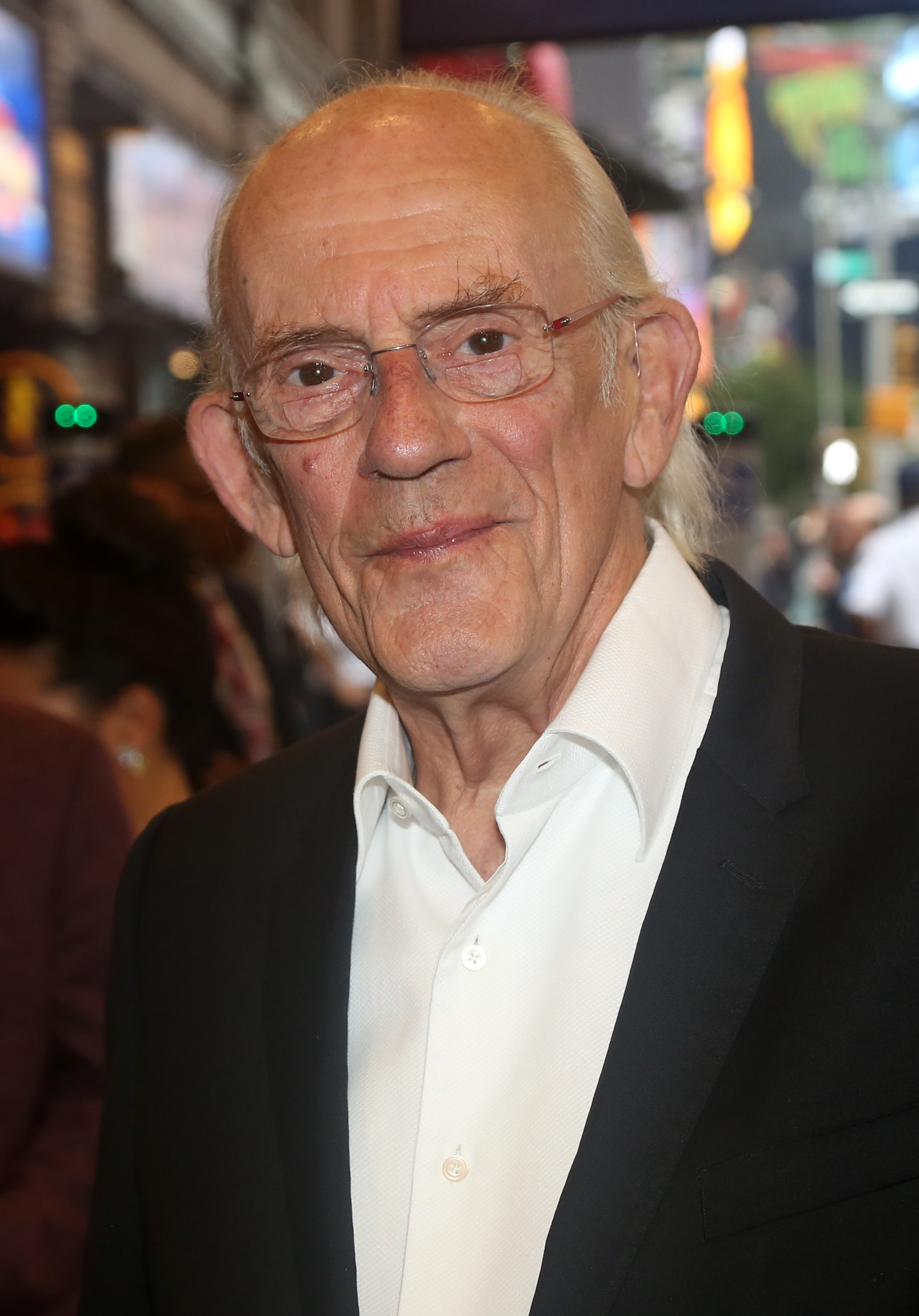 An elder man with glasses, wearing a white shirt and black blazer, smiles at a public event. The background is blurred, suggesting a bustling street or city scene