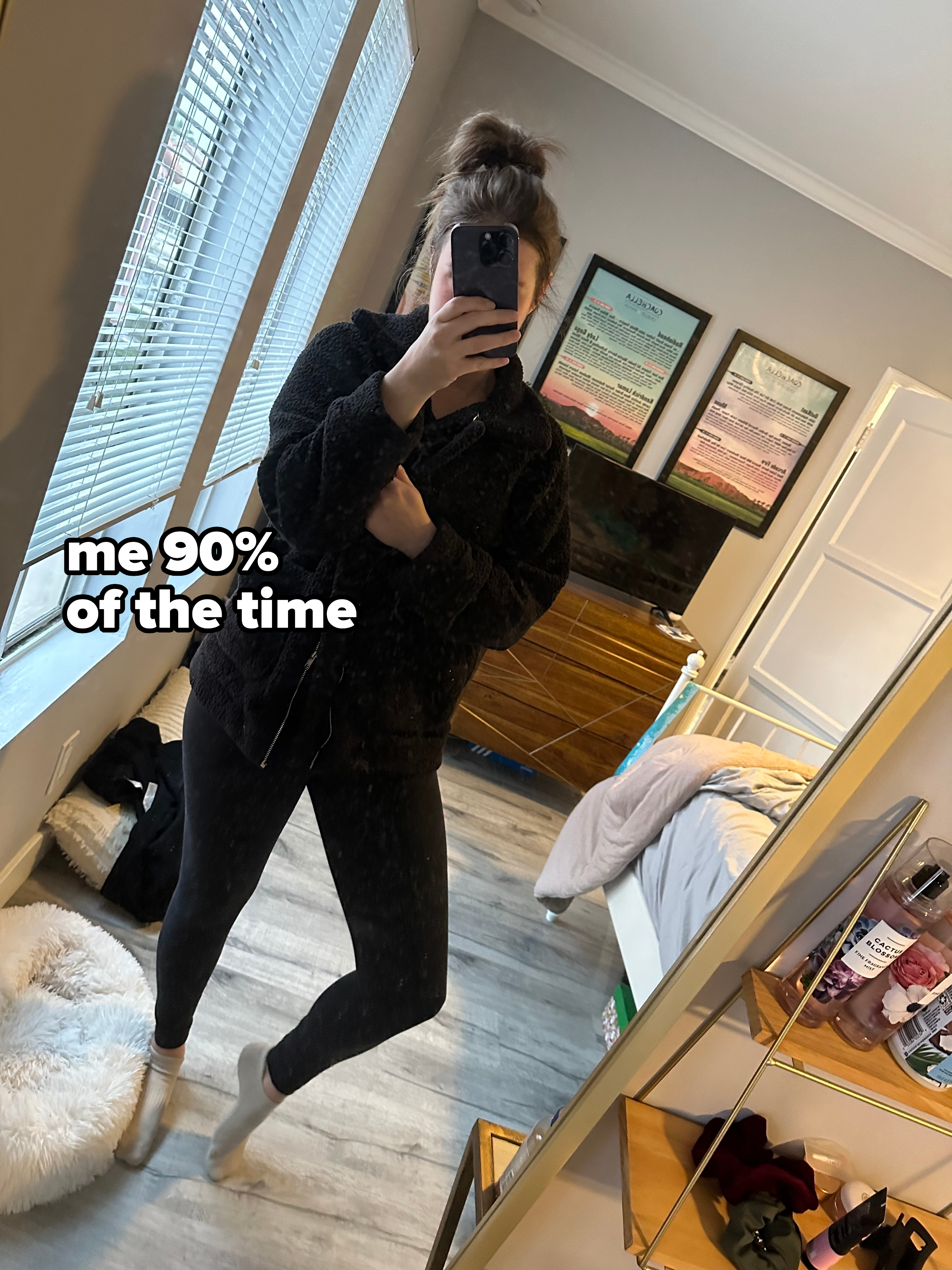 A woman takes a mirror selfie in a bedroom. She is wearing black leggings and a furry dark jacket. The room appears tidy with a bed, dresser, and framed pictures on the wall