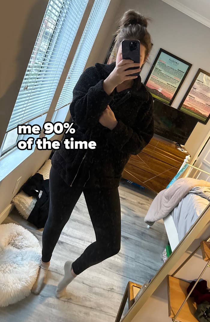 A woman takes a mirror selfie in a bedroom. She is wearing black leggings and a furry dark jacket. The room appears tidy with a bed, dresser, and framed pictures on the wall
