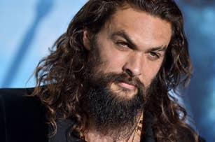 Jason Momoa at a red carpet event, wearing a black suit and sporting long wavy hair and a beard