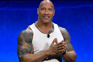 Dwayne "The Rock" Johnson stands with clasped hands, wearing a sleeveless white shirt and black pants, showcasing his tattooed arms