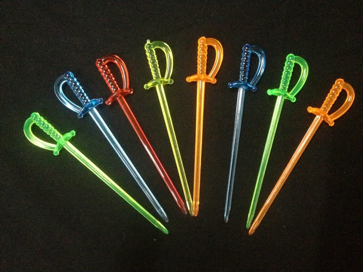Seven plastic cocktail swords are laid out side by side in a row against a black background