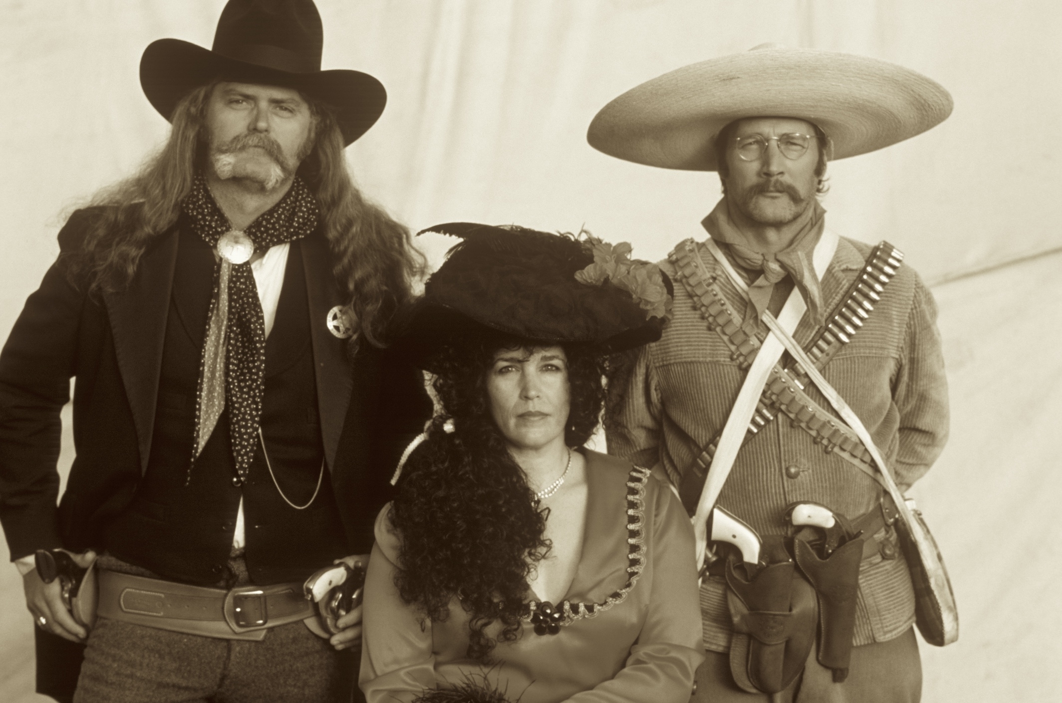 Three individuals in period Western attire are posing; two men with mustaches wearing wide-brimmed hats and gun belts, and a woman with curly hair in a dress and feathered hat