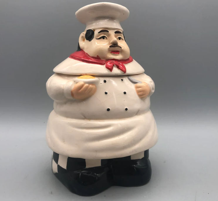 A ceramic cookie jar in the shape of a rotund chef wearing a tall hat, red scarf, and black-and-white checkered pants, holding a small item in one hand