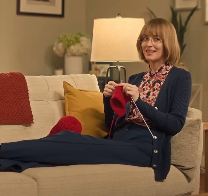 A woman with short hair, wearing glasses and a patterned blouse with a cardigan and trousers, knits on a sofa in a cozy living room