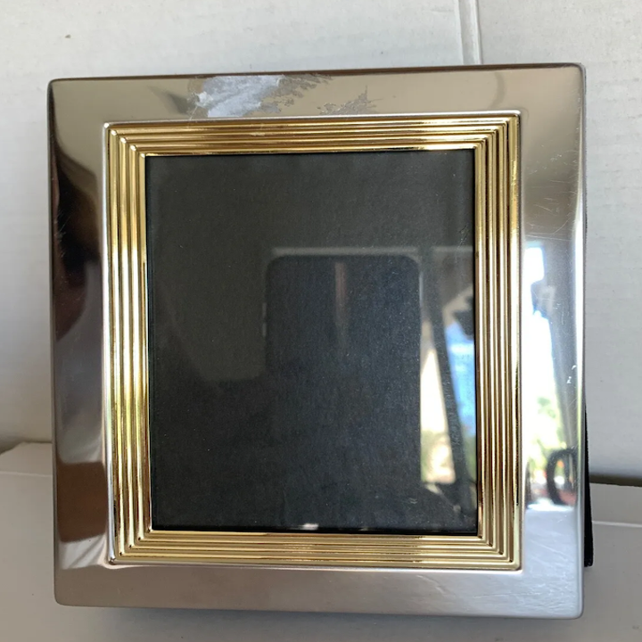A silver and gold picture frame with an empty black interior, standing upright against a neutral background