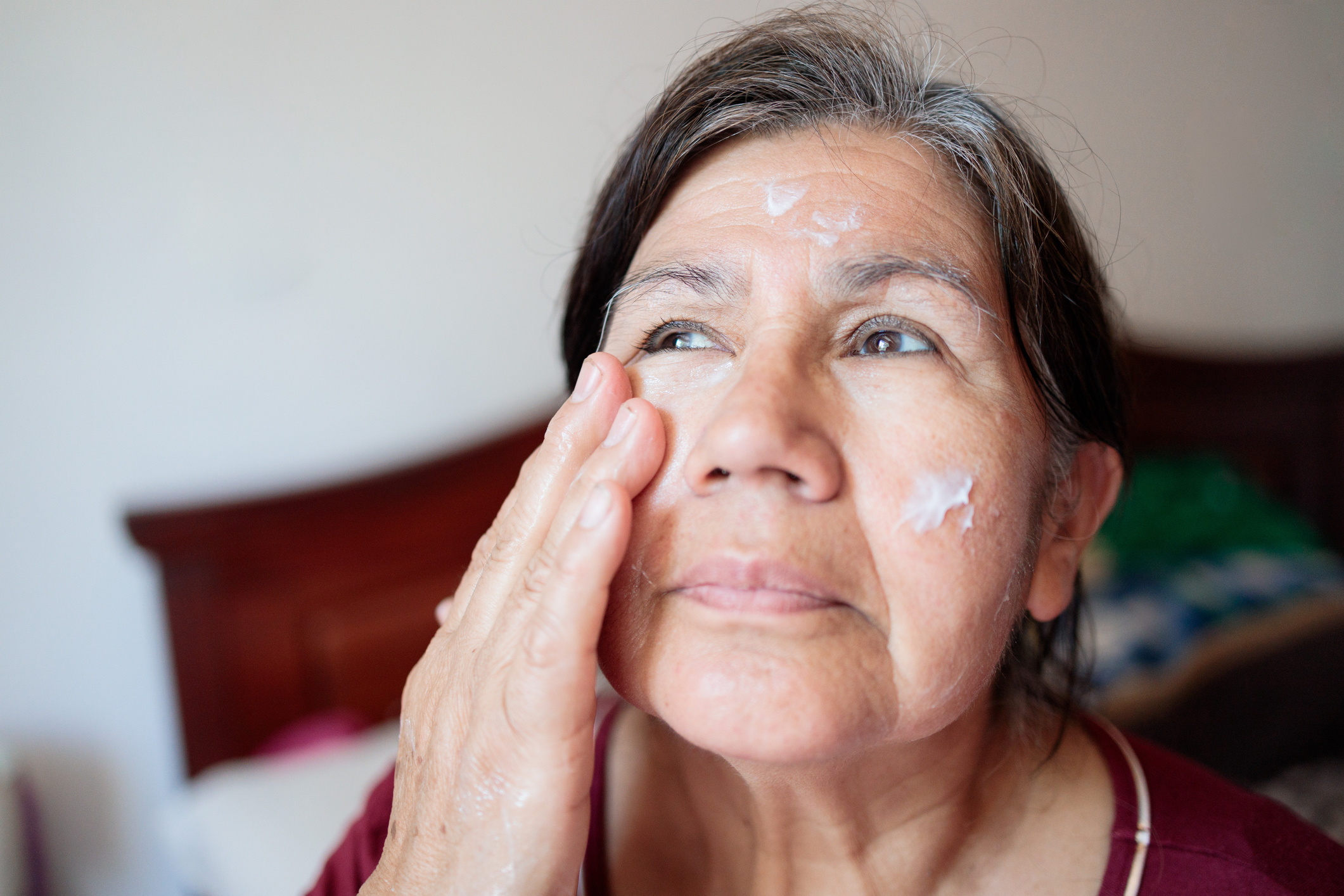 An older woman is applying lotion to her face with one hand, looking upwards with a thoughtful expression