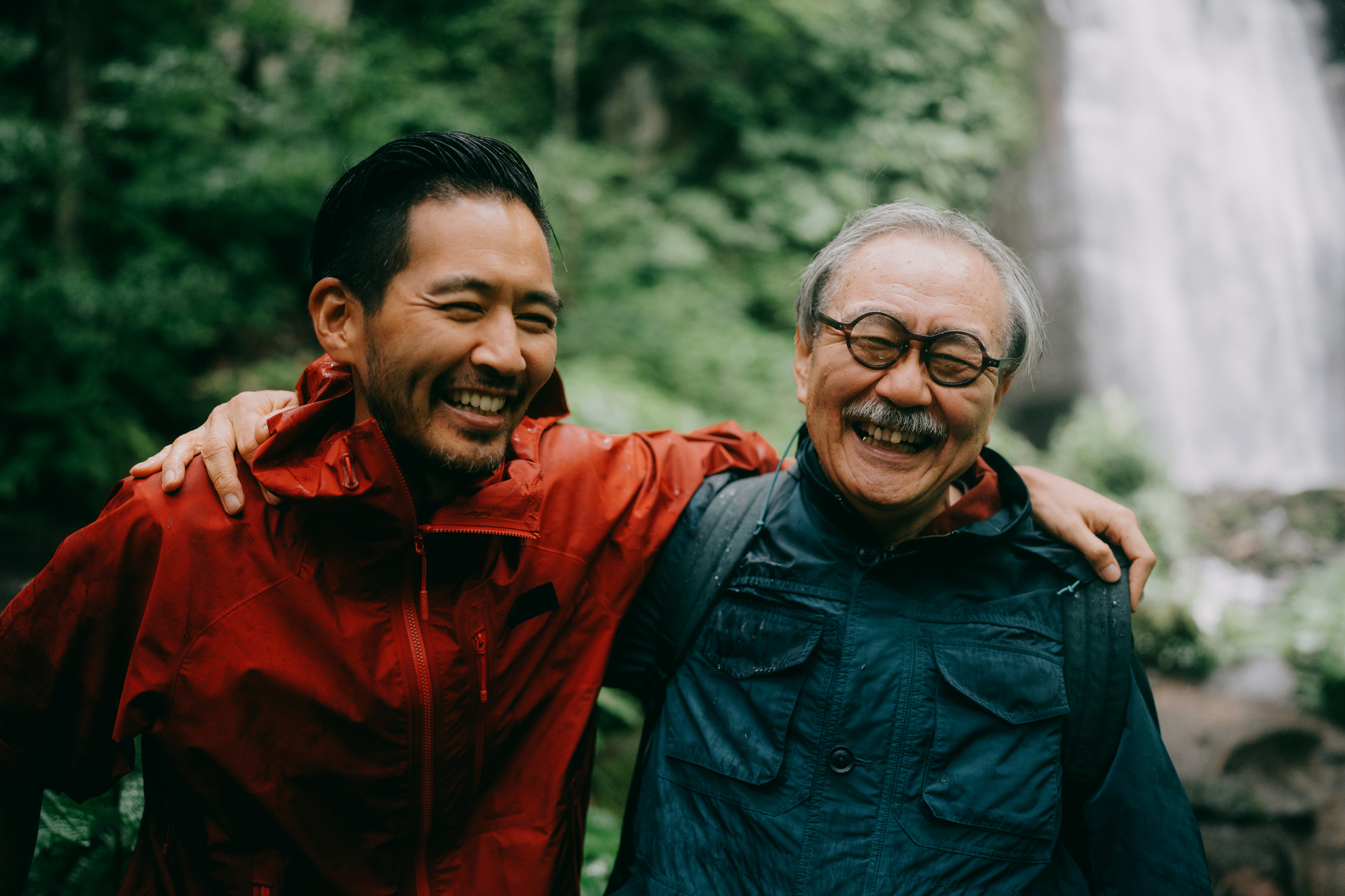 Two people, one younger and one older, smiling and wearing outdoor jackets, embrace in front of a blurred waterfall and greenery background