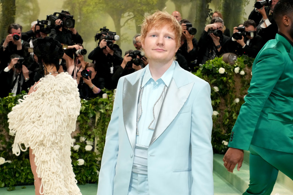 Ed Sheeran in a light-colored suit, photographed by paparazzi on a red carpet event with lush greenery in the background