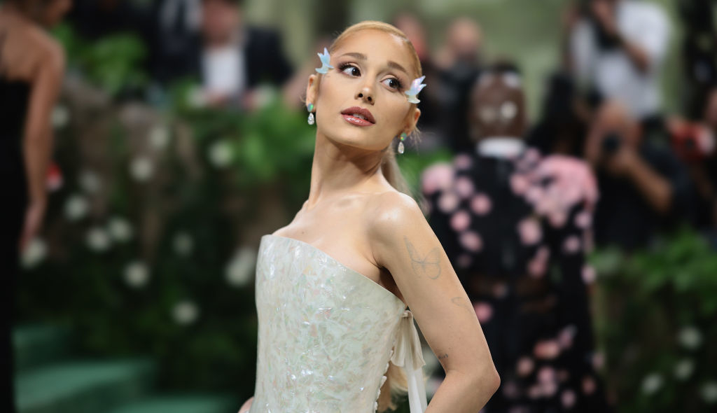 Ariana Grande on a red carpet wearing an elegant strapless gown with floral accents. She is adorned with flower-shaped accessories in her hair
