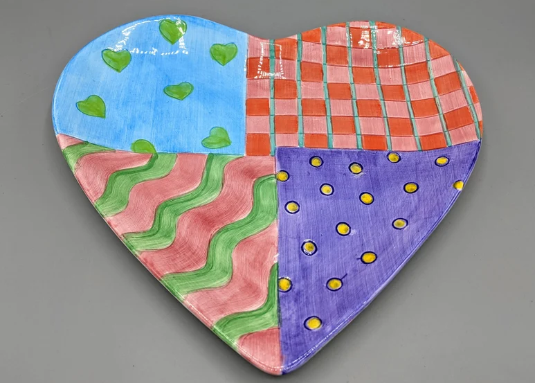Heart-shaped ceramic plate with four quadrants, each with distinct patterns: green hearts on blue, red checkered, wavy green lines on pink, and yellow dots on purple