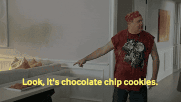A man in a red bandana and a graphic t-shirt points to a plate of cookies on a table, saying, "Look, it's chocolate chip cookies."