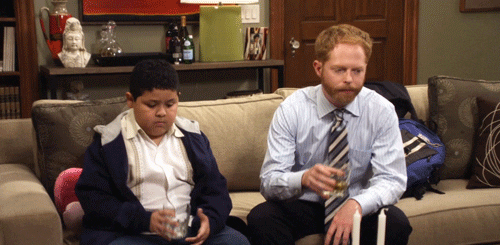 Rico Rodriguez and Jesse Tyler Ferguson sit on a couch holding drinks, looking downcast. The background shows a decorated living room