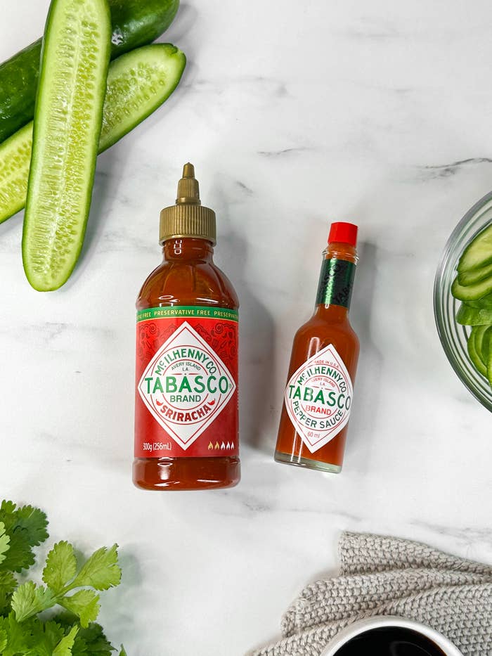Two Tabasco sauce bottles, one green and larger, the other red and smaller, surrounded by sliced cucumbers, cilantro, and a dish of cucumbers