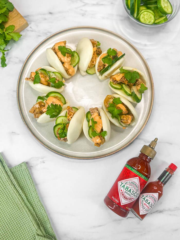 Plate of bao buns with crispy chicken, cucumber slices, cilantro, and drizzled sauce, next to bottles of Tabasco hot sauce and a green napkin