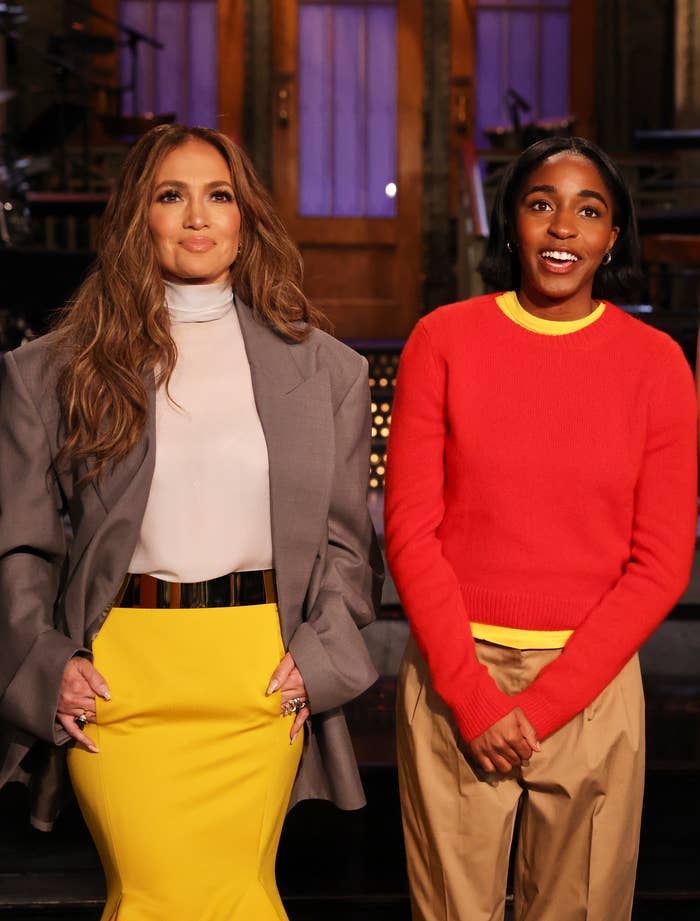 Jennifer Lopez, Ayo Edebiri stand together on a set. Jennifer wears a gray blazer over a white top and yellow skirt, Ayo wears a red sweater and khakis.