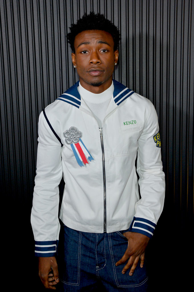 Niles Fitch wearing a Kenzo jacket and dark pants poses in front of a vertical striped background
