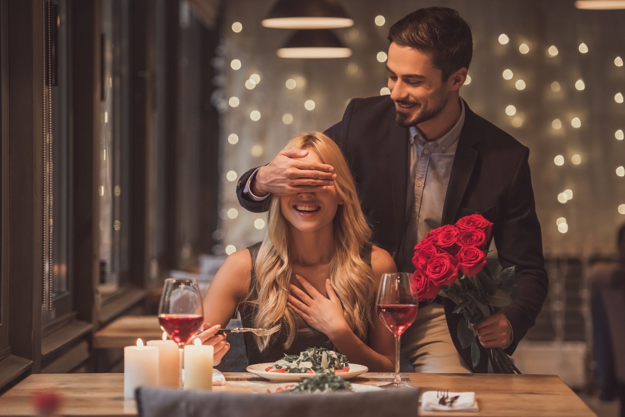 A man covers a smiling woman's eyes with one hand while holding a bouquet of red roses in the other