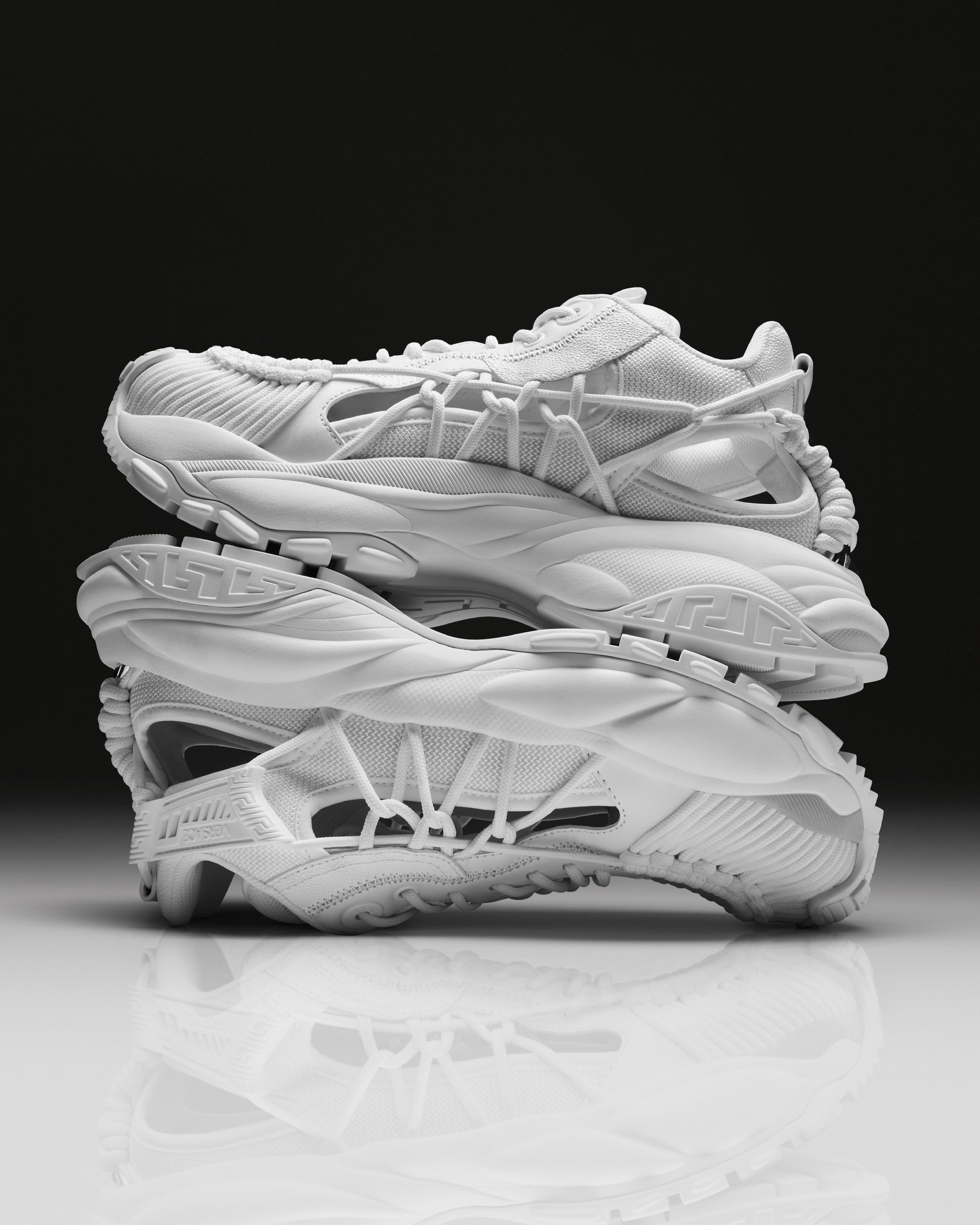 Stack of three white, intricate, modern sneakers with detailed textures and futuristic design elements, displayed against a black background. No people present