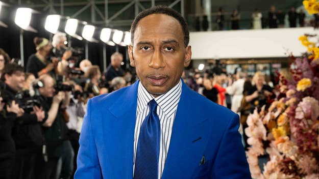 Stephen A. Smith is shown on a red carpet, dressed in a blue suit with a striped shirt and a dark tie. Photographers and cameras are in the background