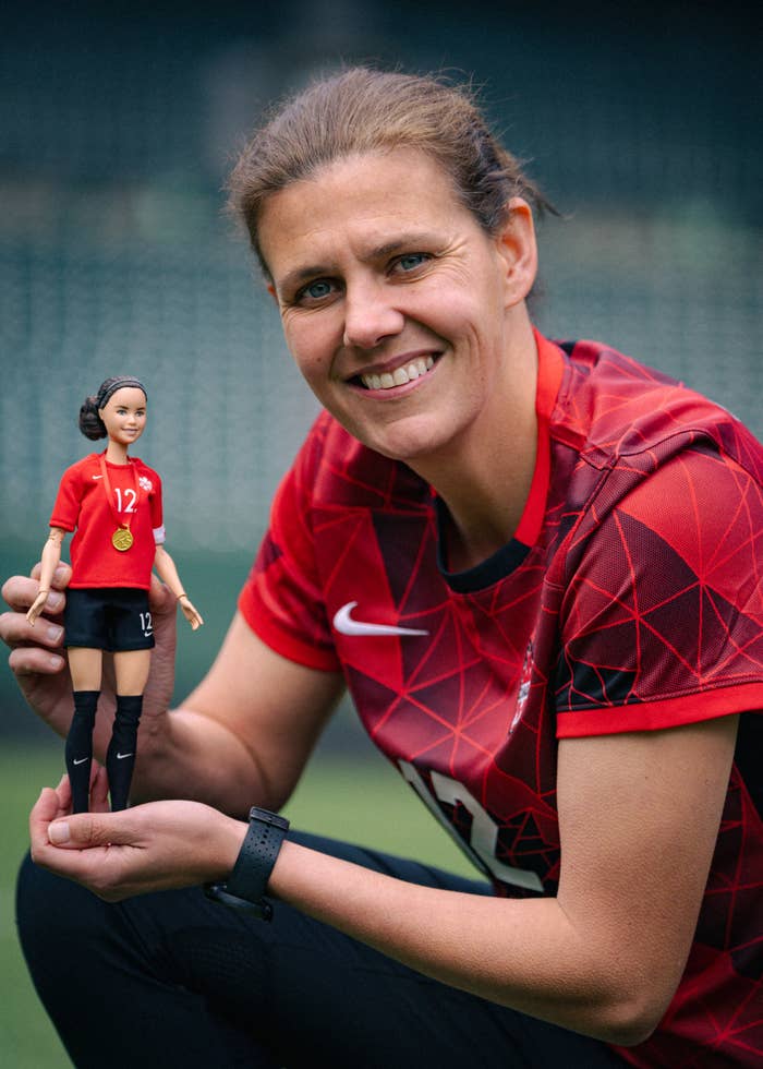 A woman smiles, holding a doll wearing a matching soccer uniform. The woman, whose shirt has the number 12, appears to be a soccer player