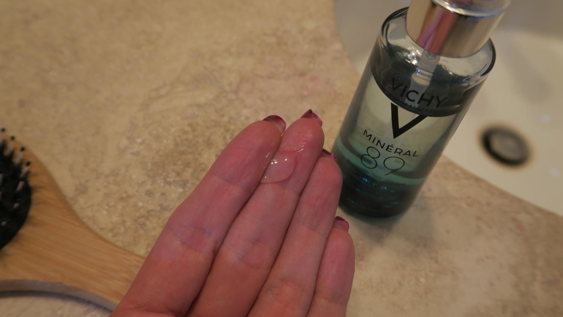 A Close-up of a reviewer's hand displaying Vichy Mineral 89 serum in a bathroom. A hairbrush is partially visible in the background