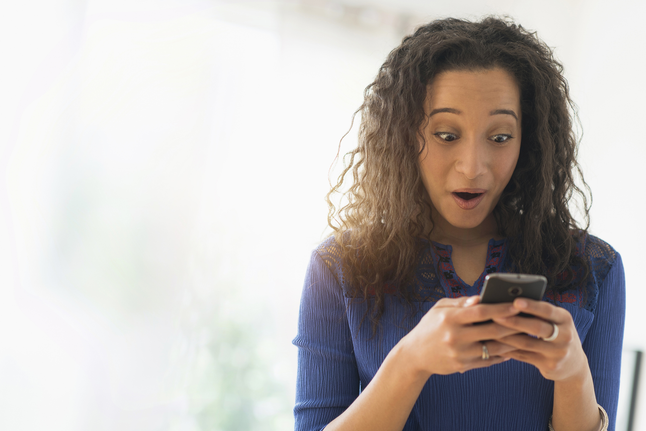 A woman with curly hair looks at her phone in surprise, seemingly astonished or excited by what she is seeing