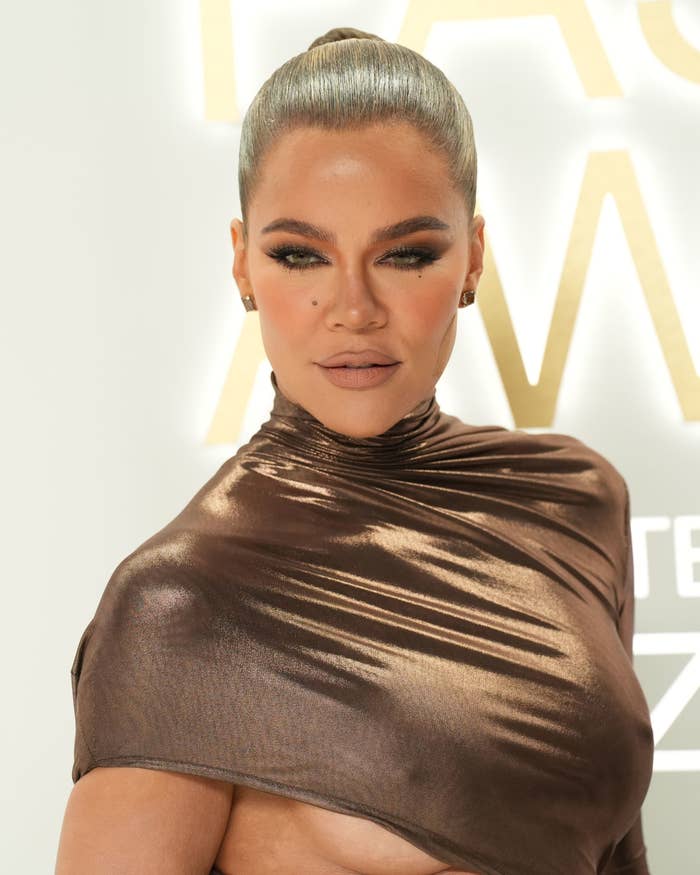 Khloé Kardashian at an event, wearing a metallic off-shoulder dress with sleeked-back hair