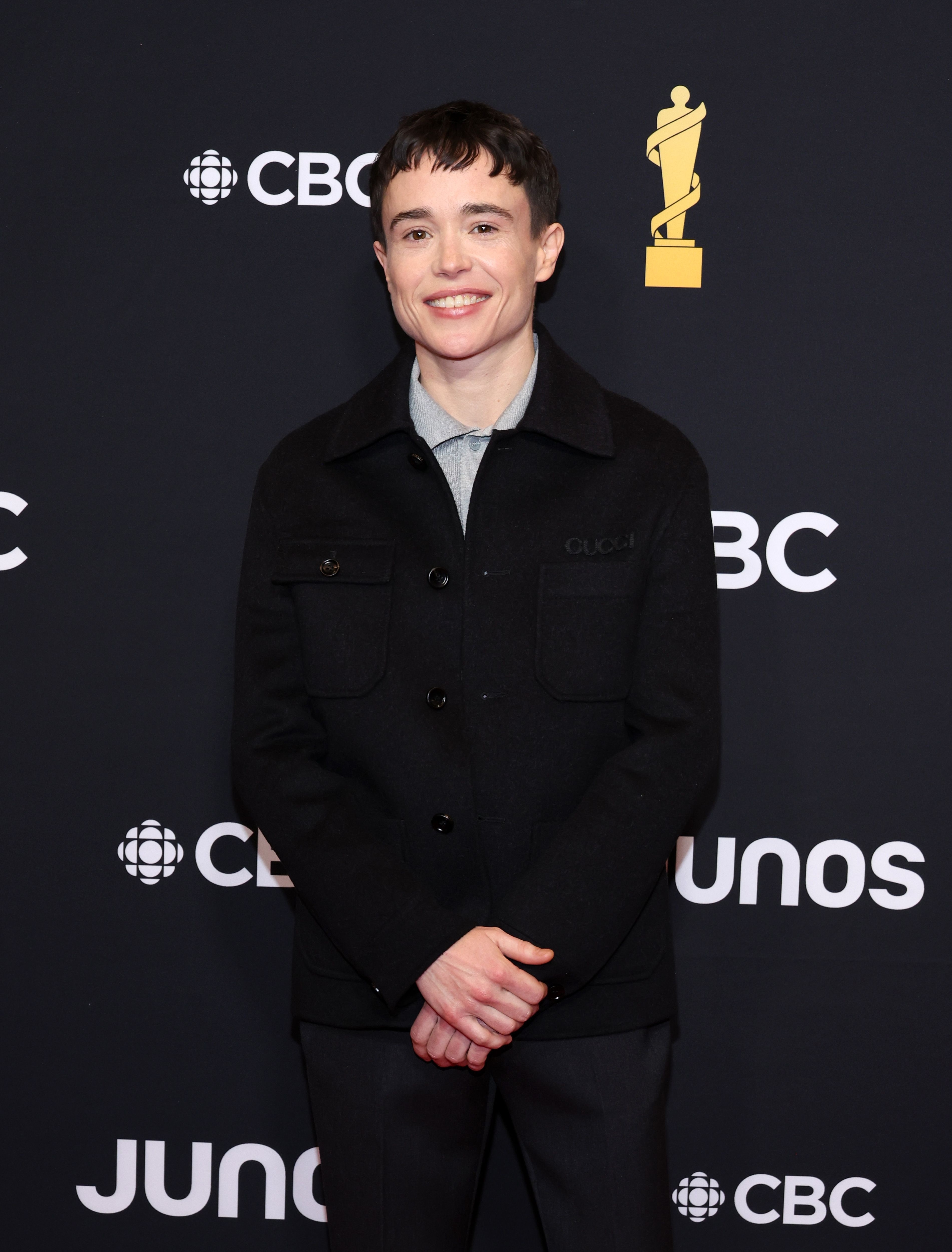 Elliot Page on the red carpet at the Juno Awards, wearing a black jacket and dark pants, standing and smiling at the camera