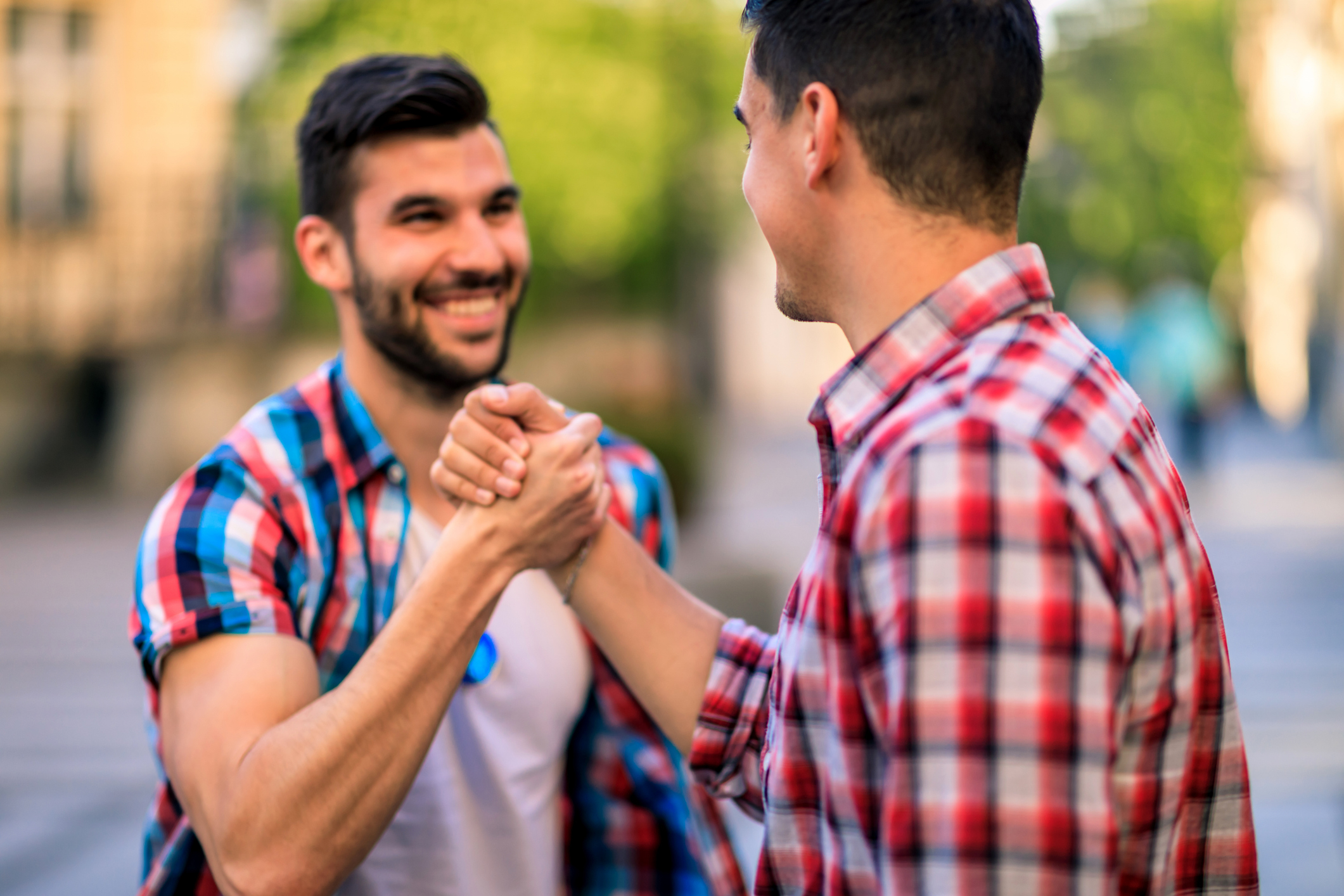 Two men wearing casual plaid shirts greet each other with a friendly handshake outdoors, both smiling warmly
