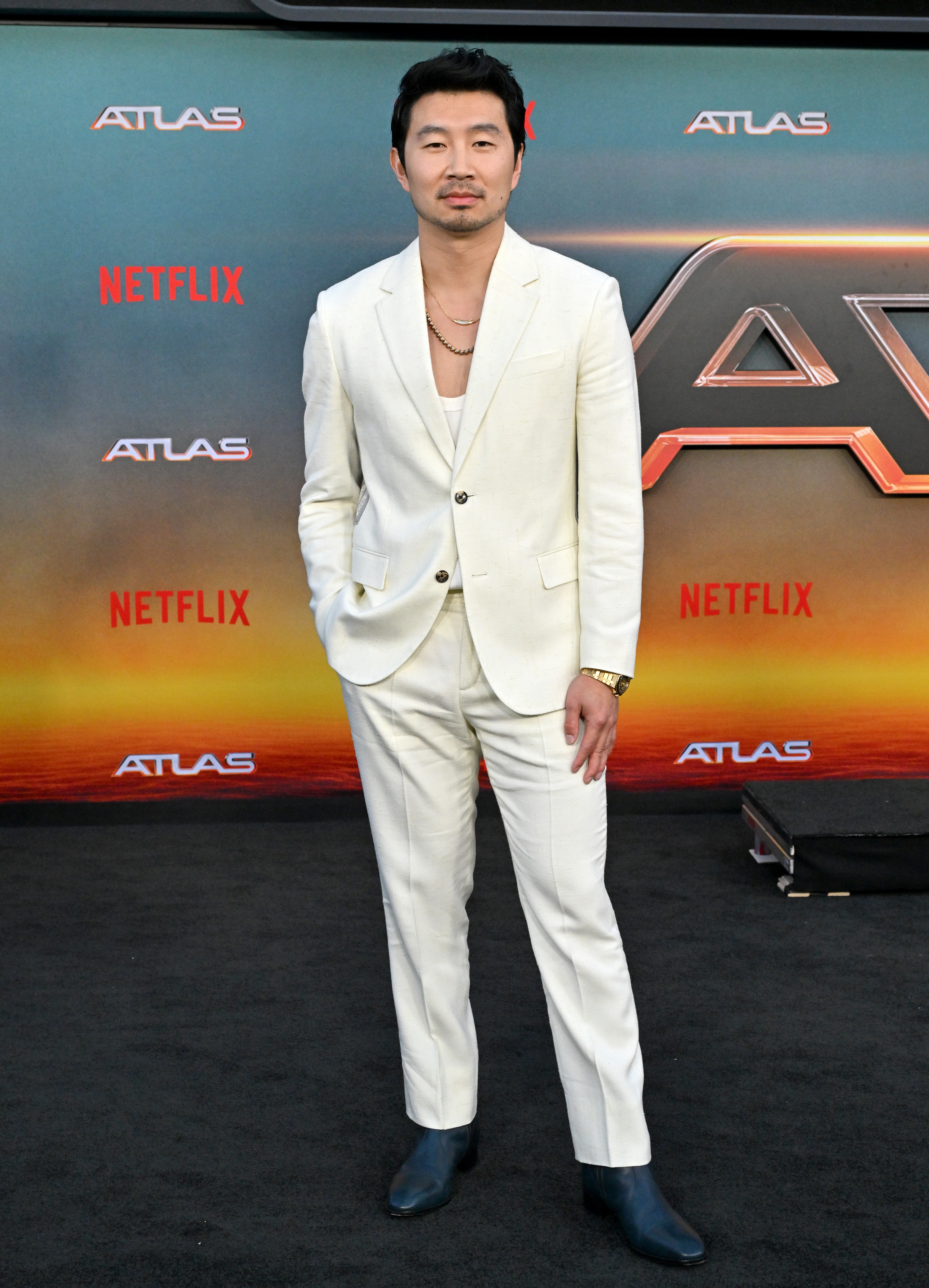 Simu Liu poses on the red carpet in a white suit, gold watch, and black shoes at a Netflix "ATLAS" event