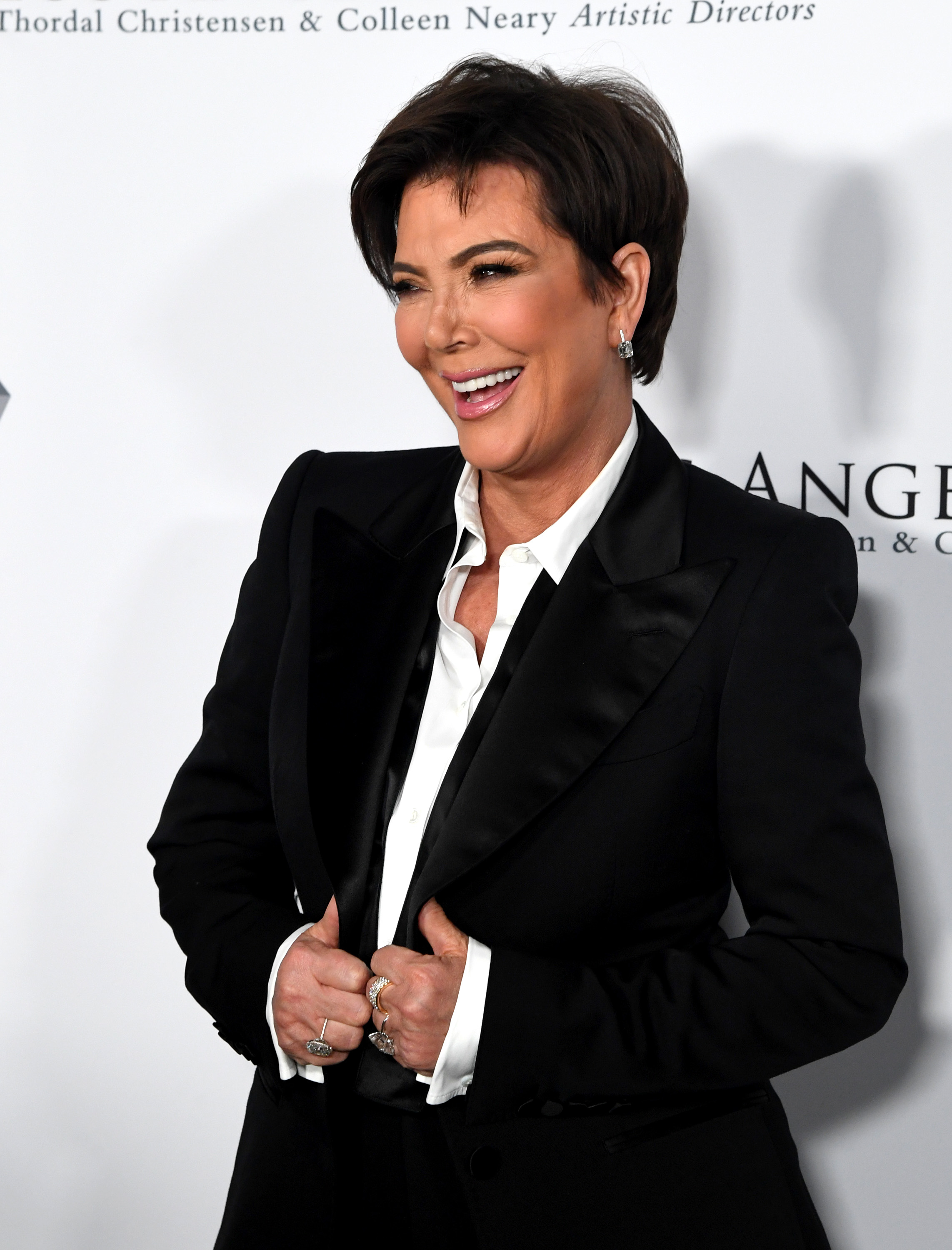 Kris Jenner is smiling on the red carpet, wearing a black tuxedo-style outfit with a white collared shirt. She is buttoning her jacket and wearing rings