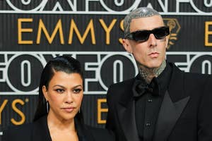 Kourtney Kardashian and Travis Barker on a red carpet, both wearing black outfits. Kourtney is in a blazer and gloves, and Travis is in a suit with sunglasses