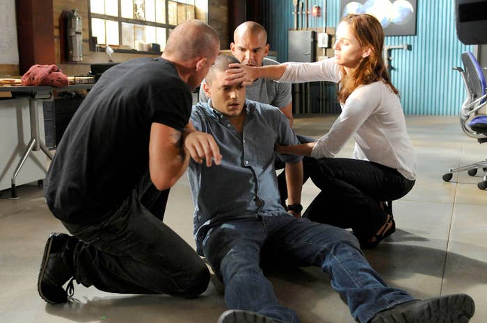 A group of individuals is assisting Wentworth Miller, who appears to be unwell, lying on the floor in a scene from Prison Break