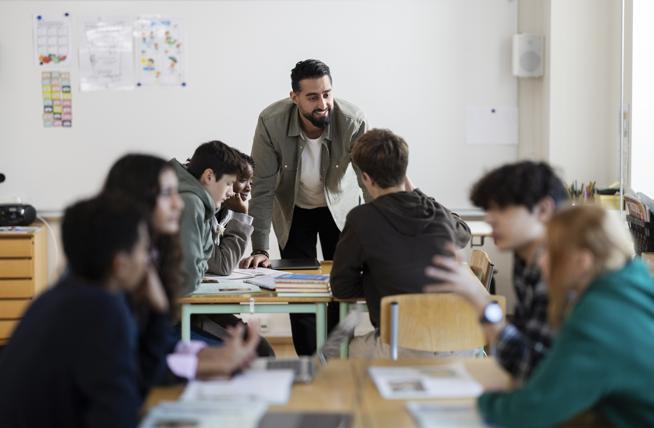A teacher in casual attire engages with high school students seated around a table in a classroom. Various educational materials are on the tables